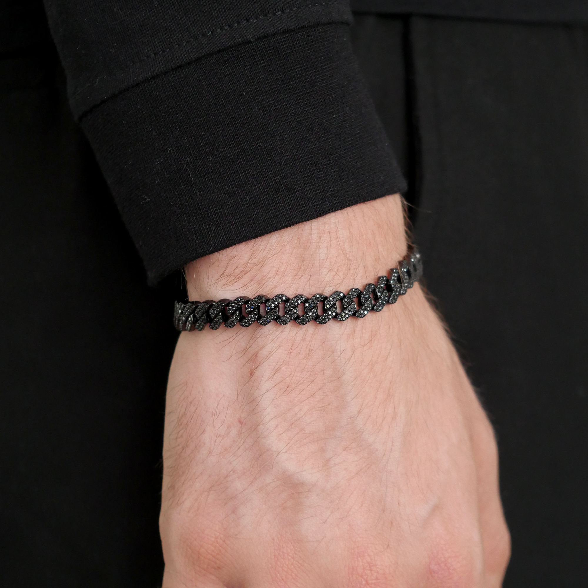 A close-up of a person's wrist wearing a black chain bracelet with sparkling dark stones, set against a black long-sleeve shirt.