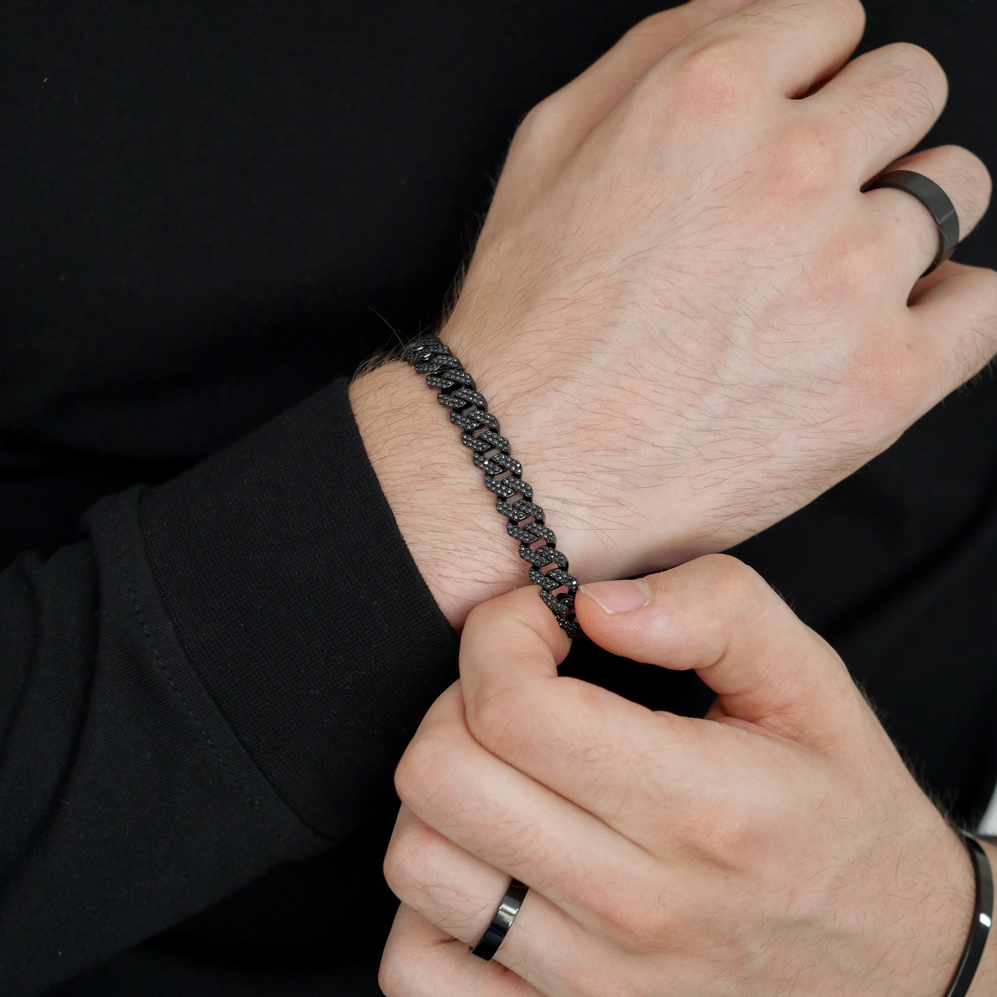 A close-up of a man's wrist wearing a black frosted cuban bracelet, with his hands adjusting the strap. He is wearing a black long-sleeve shirt and black rings on his fingers.